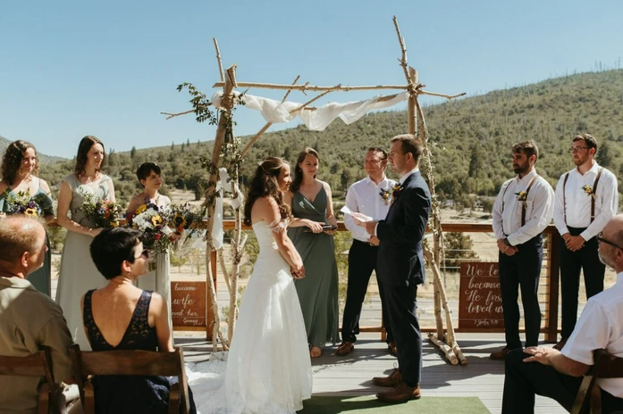 A wedding ceremony outdoors, with a couple under a wooden arch, surrounded by their wedding party and guests seated nearby.