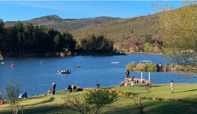 People by a lake with boats, surrounded by trees and hills, under a clear blue sky.