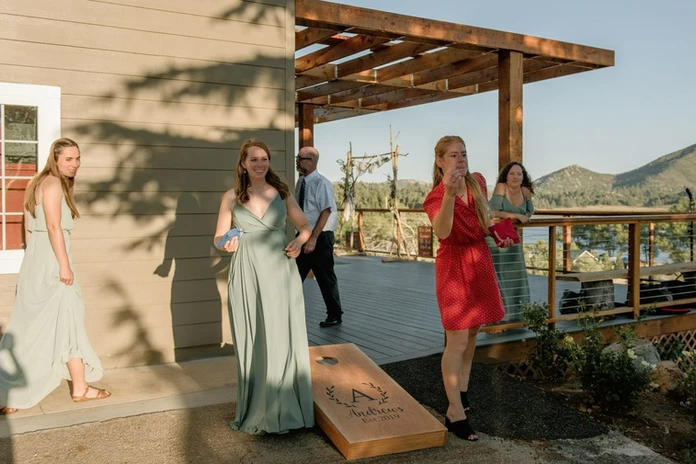 People outdoors playing cornhole, some in dresses, one in red. A wooden pergola and scenic view in the background.