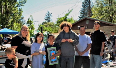 A group of people standing outdoors, some holding a box with a tablet or gadget image on it. Trees and a building in the background.