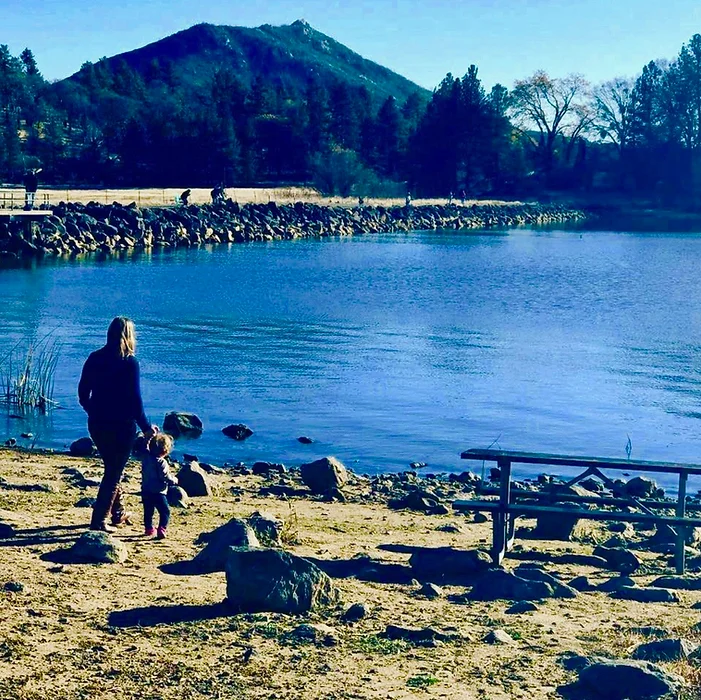 A person and a child walking along a lake shore with a mountain and trees in the background.