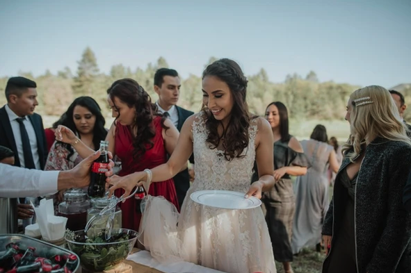 People in formal attire at an outdoor event, gathering food and drinks from a table.