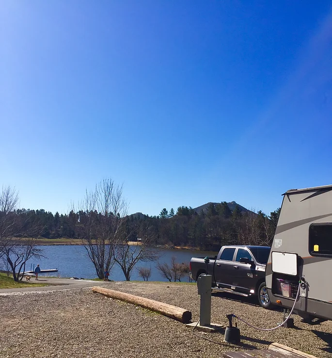 A pickup truck and RV near a lake, with trees and hills in the background under a clear blue sky.