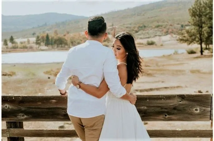A couple embraces on a wooden fence, overlooking a scenic landscape with hills and a lake.