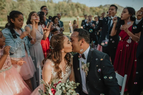 A couple kissing surrounded by people blowing bubbles, dressed in formal attire at an outdoor event.