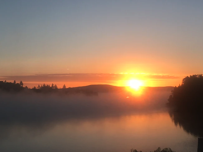 Sunrise over a misty lake, with silhouetted trees and hills in the background, reflecting warm orange and yellow hues on the water.