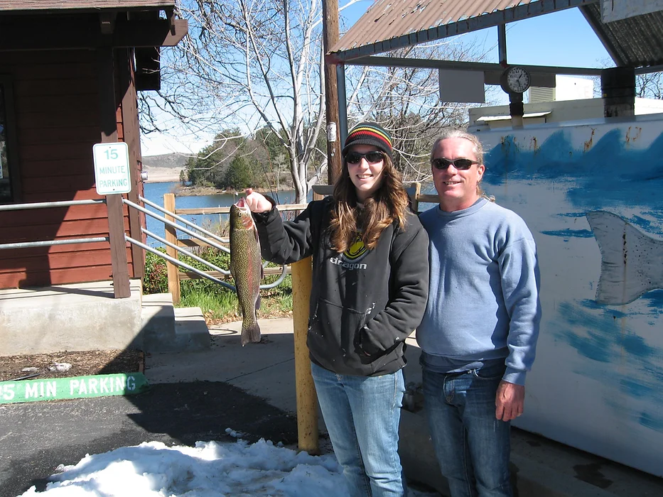 Two people outdoors, one holding a fish, posing for a photo by a building with a "15 Minute Parking" sign nearby.