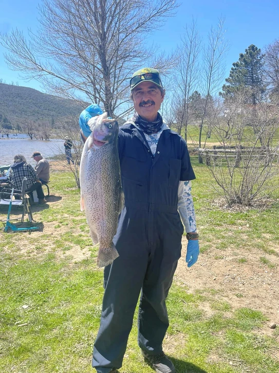 Person holding a large fish outdoors near a lake, with trees and people sitting nearby.