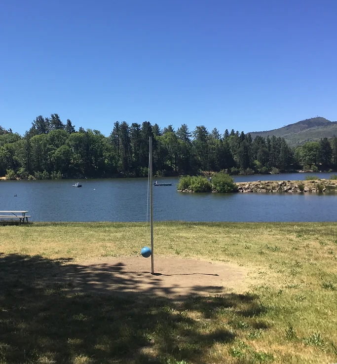 A tetherball pole and ball on grass near a serene lake with trees and a clear blue sky in the background.