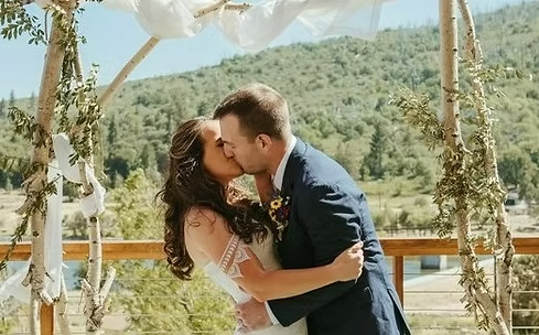 A couple kissing under a rustic wedding arch with a scenic outdoor background.