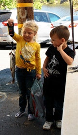 Two children with a fish; one holds it up, smiling, while the other looks on. Cars are parked in the background.