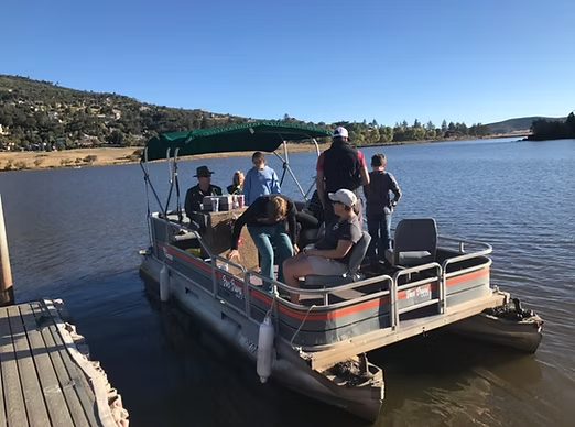 A group of people are on a pontoon boat on a lake, with hills and trees in the background.