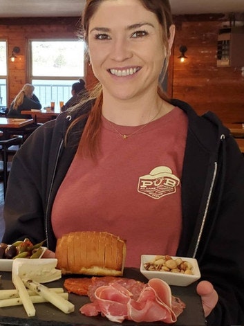 A person holding a platter with various cheeses, meats, bread, and olives in a cozy indoor setting.