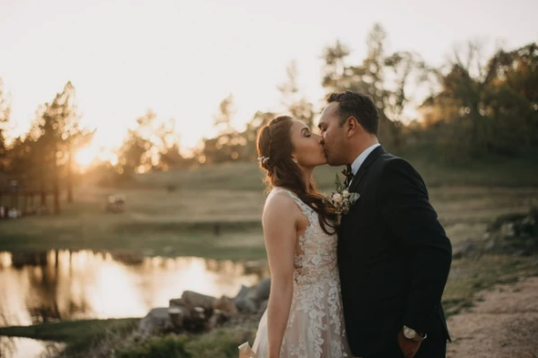 A couple kissing at sunset, dressed in wedding attire by a pond with trees in the background.