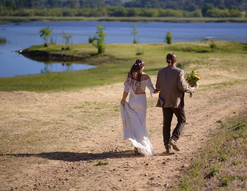 A couple in wedding attire walking by a lake holding a bouquet.