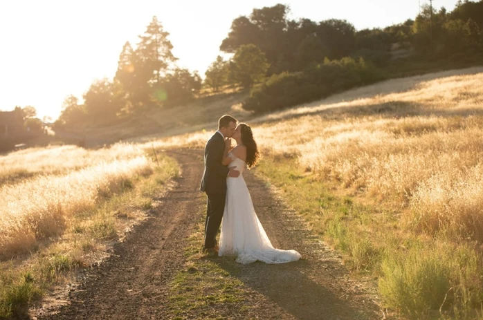 A couple in wedding attire kisses on a sunlit dirt path in a scenic field.