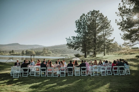 Outdoor gathering by a lake, guests seated at tables, surrounded by trees and mountains under a clear sky.