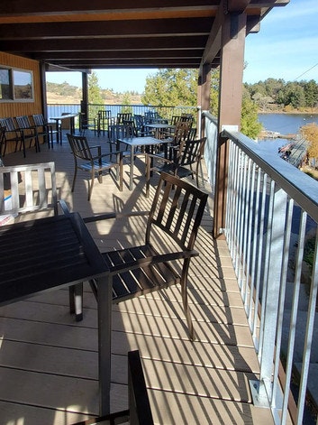 Outdoor patio with empty black metal tables and chairs overlooking a scenic lake and trees.