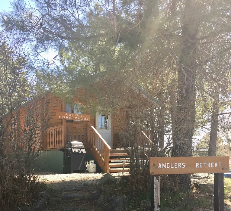 A wooden cabin in a forested area, with a sign reading "Anglers Retreat" and "Guest Cabin Please Do Not Disturb."