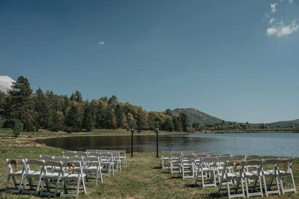 An outdoor ceremony setup by a lake with rows of white chairs, surrounded by trees and mountains under a clear blue sky.