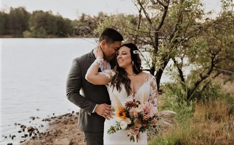 A bride and groom pose by a lakeside, surrounded by trees. The bride holds a bouquet of flowers.