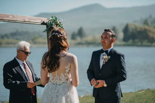 A couple is getting married outdoors by a lake, with an officiant in a suit presiding over the ceremony.