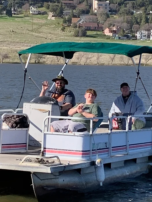 Three people are on a pontoon boat on a lake, with one person waving.