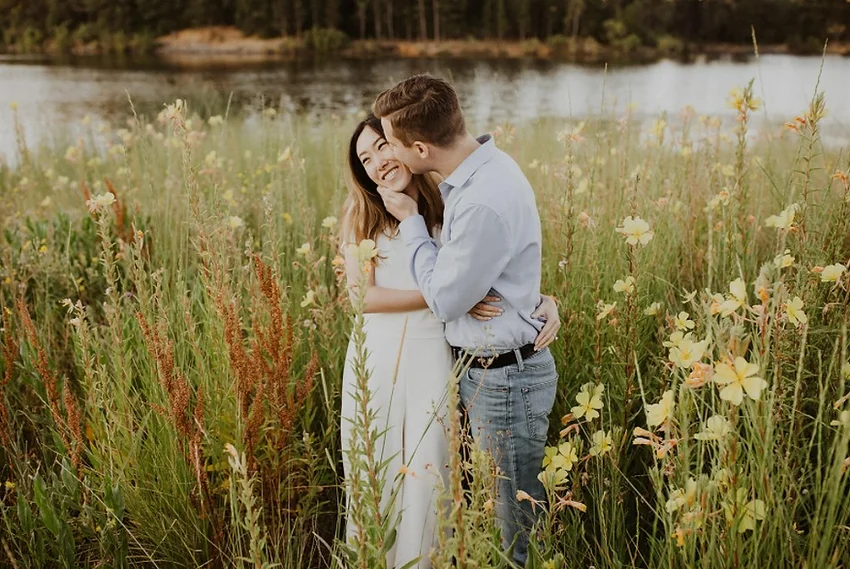 A couple embracing in a field of tall grass and yellow flowers by a lake.