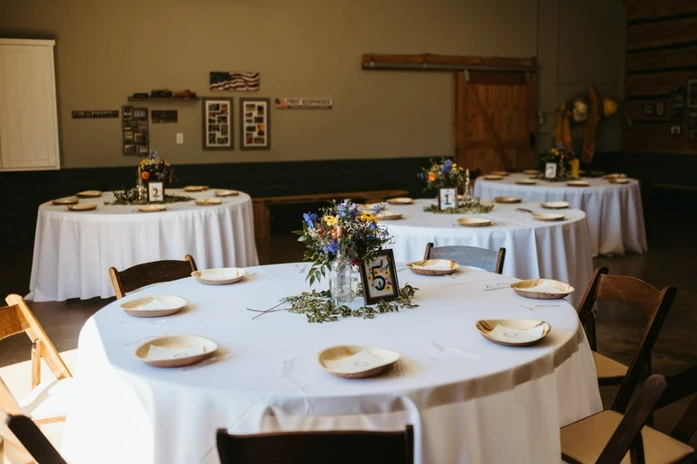 Tables set for an event with white tablecloths, wooden chairs, floral centerpieces, and plates.