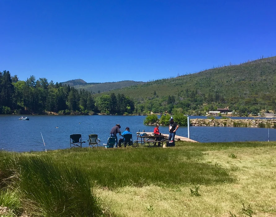 People sitting by a lakeside, with fishing chairs, a picnic table, and scenic hills in the background.