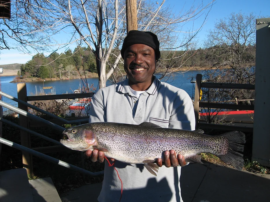 A person smiling, holding a large fish near a lake with trees and clear sky in the background.