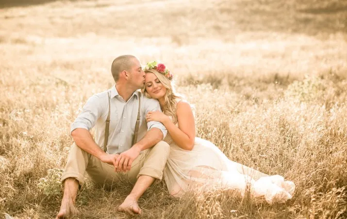 A couple sits in a field; the man kisses the woman's forehead, and she wears a flower crown and leans on him.