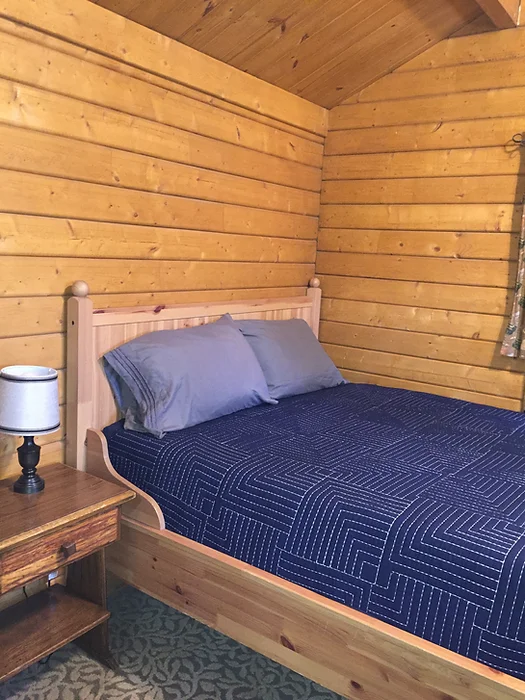 A cozy wooden bedroom with a bed, blue bedding, a side table, and a lamp.