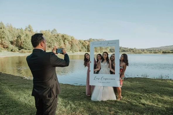 Four women pose with a photo frame near a lake while a man takes their picture.