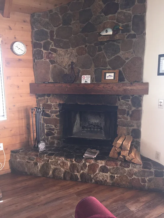 A rustic stone fireplace with a wooden mantel, clock, fire tools, decor items, logs, and a framed picture.
