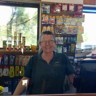 A person in a store stands behind a counter with various products displayed on shelves.