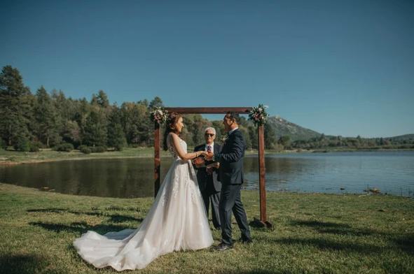 A couple is getting married outdoors by a lake, with a person officiating the ceremony under a wooden arch adorned with flowers.