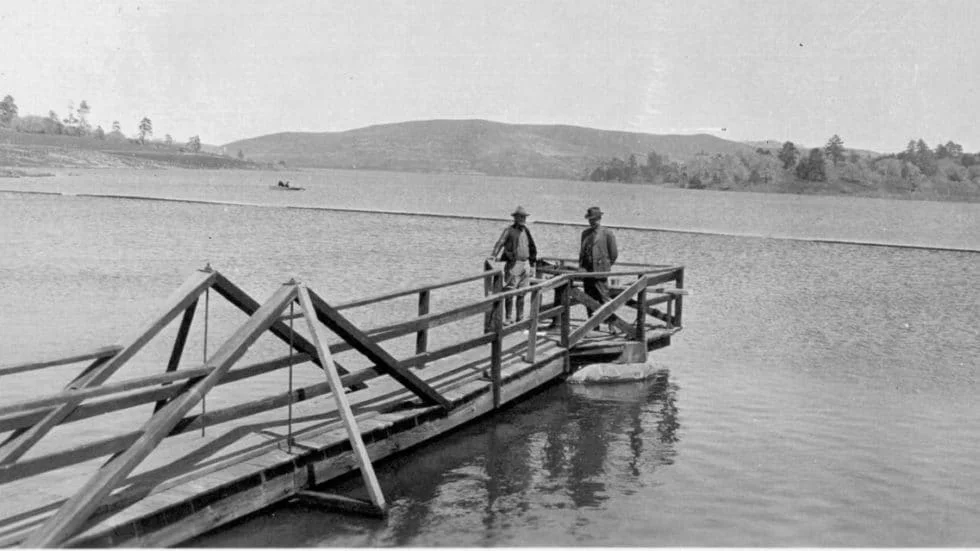 Two men stand on a dock over a lake with hills and trees in the background.