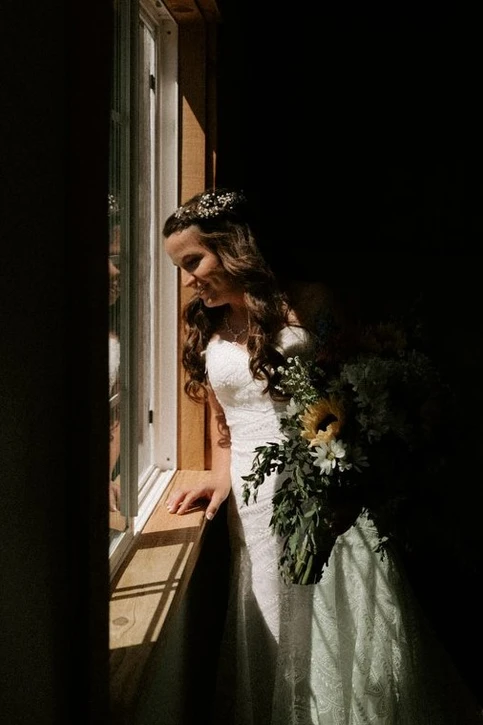 A bride in a white dress and a flower crown stands by a window holding a bouquet, gazing outside.