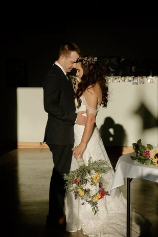 A couple stands embraced at a wedding, with the bride holding a bouquet and light casting shadows on them.
