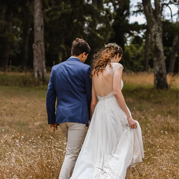 A couple in wedding attire walking hand-in-hand through a forest clearing.