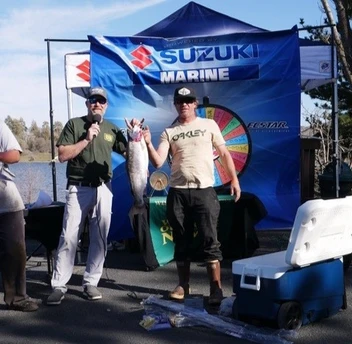 Two men at a fishing event; one is holding a large fish. There's a "Suzuki Marine" banner and a cooler nearby.