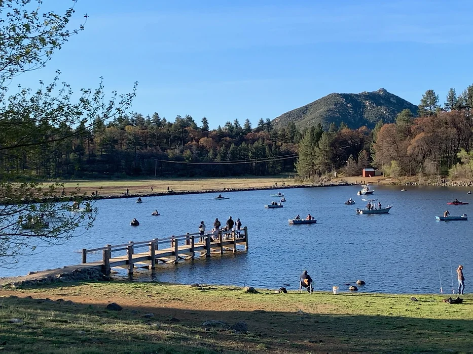 People fishing from boats and a dock on a lake, with a mountain and trees in the background.