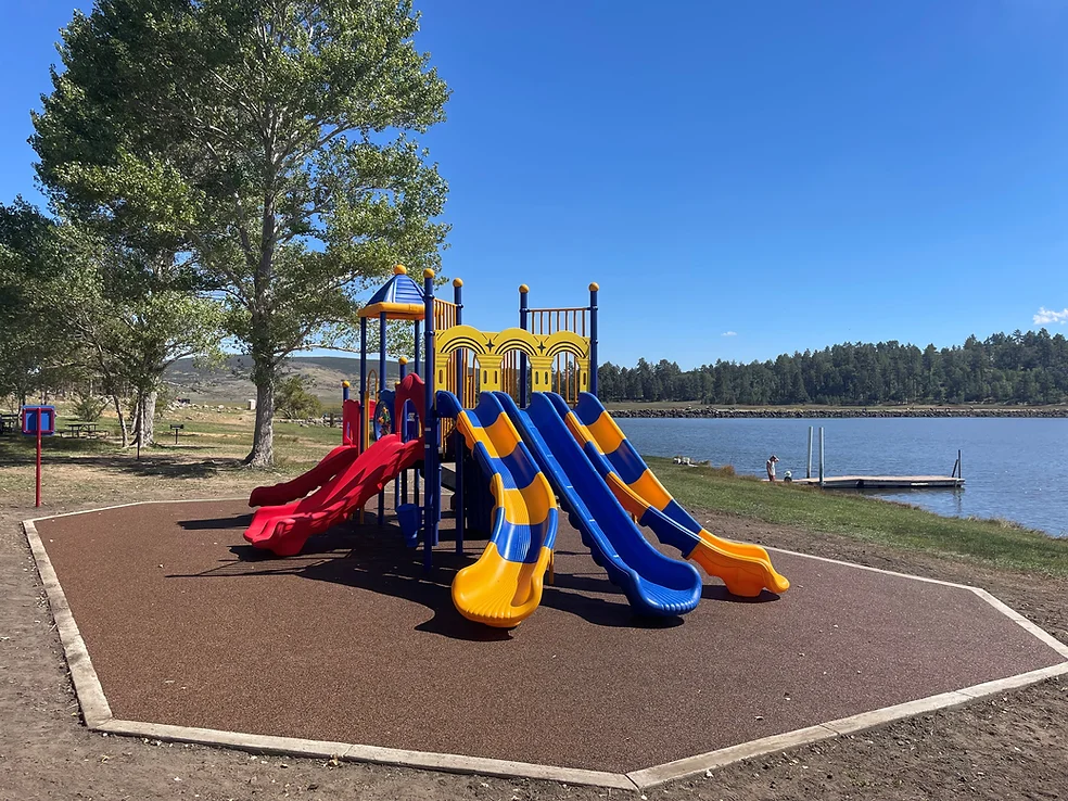 A colorful playground with slides near a lake, surrounded by trees and a clear blue sky.