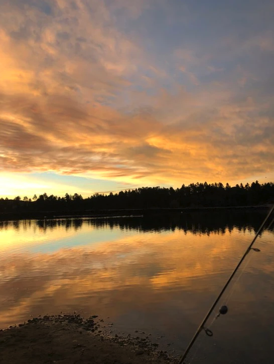 A serene lake at sunrise or sunset with a fishing rod, reflecting vibrant orange clouds and silhouetted trees.