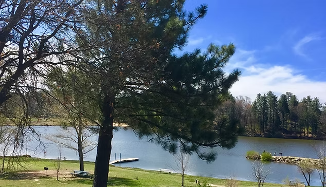 A serene lake scene with trees, a dock, and grassy areas under a clear blue sky.