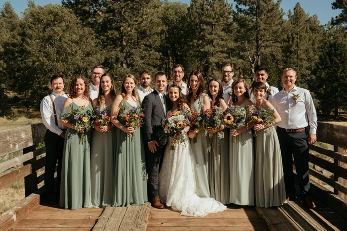 A wedding party poses outdoors, featuring bridesmaids in green dresses and white-shirted groomsmen, with trees in the background.
