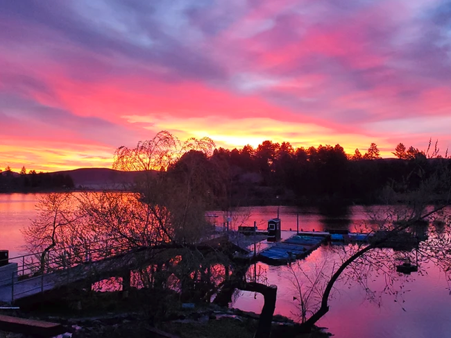 A vibrant sunset over a lake with silhouetted trees and a dock.