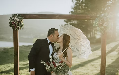 A couple kissing under a wooden arch with floral decorations, holding an umbrella and bouquet, in an outdoor setting.