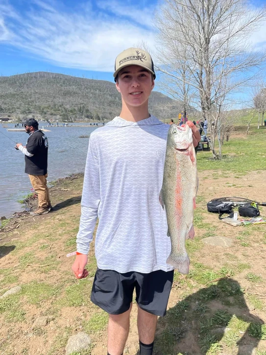 A person in a cap holds a large fish by a lake. Another person fishes in the background.